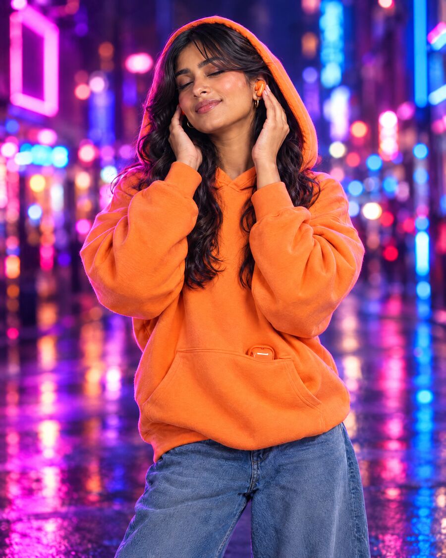Model in a tangerine hoodie with the hood up on a neon-lit street at night